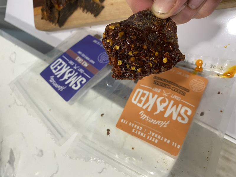 Close-up of a thick, chili-crusted piece of beef jerky with a branded pouch behind.