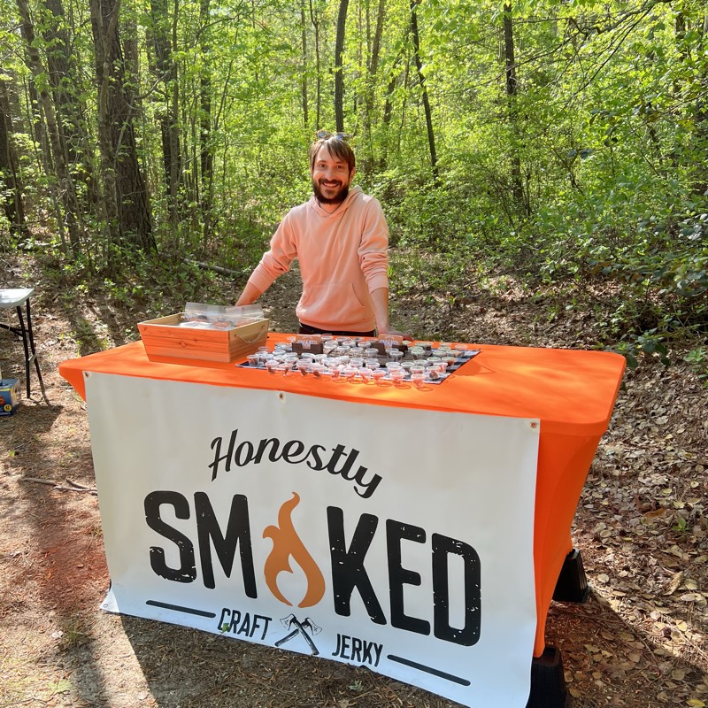 Founder behind an Honestly Smoked branded booth with jerky sample cups in a sunlit forest.