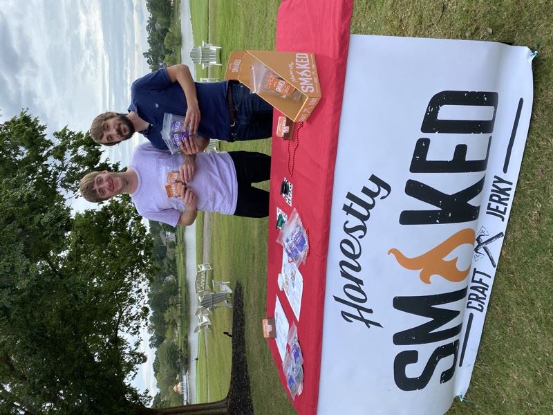 Two founders with jerky bags behind the Honestly Smoked banner table by a lake.